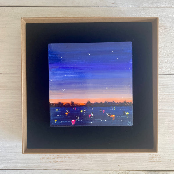 Boxed artwork of a sunset over water with colorful tow floats, on a wooden surface.