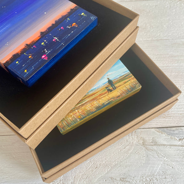 Two rectangular paintings inside an open cardboard box stacked on a light wooden surface.