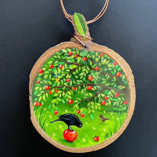 A circular wooden slice miniature painting featuring an apple tree with red apples, a black bird pecking at an apple, and a copper wire hanging loop at the top, shown against a dark background.