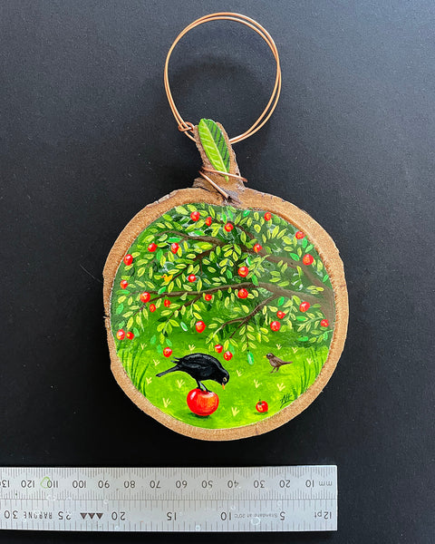 A round hand-painted wooden slice painting featuring a green apple tree with red apples and a black bird eating an apple, suspended from a copper wire loop. A metal ruler is placed below the ornament to show scale against a dark background.