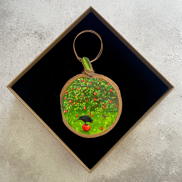 A miniature painting on wood slice featuring an apple tree scene with green leaves, red apples, and a black bird, displayed inside a black square gift box on a textured light gray surface.