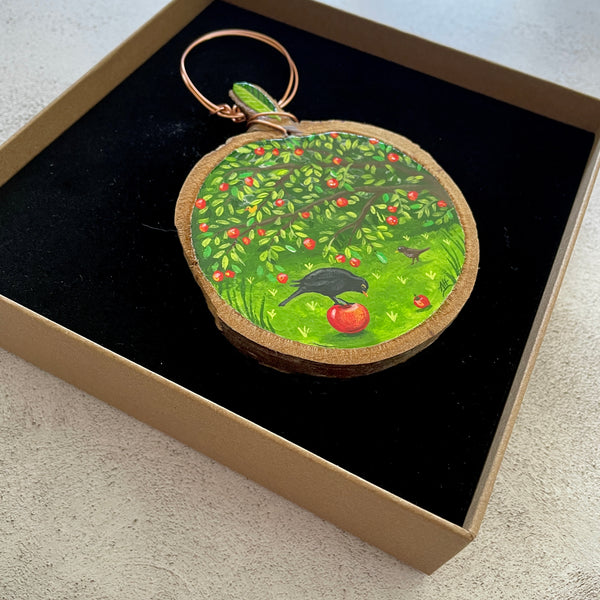 A round wooden slice miniature painting featuring a painted green apple tree with red apples and a black bird, displayed inside a brown gift box with a black velvet lining.
