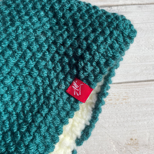 A close-up of a teal green chunky knit headband with a soft cream sherpa lining and a small red fabric tag featuring a white embroidered Andrea Hall logo, laid flat on a light wood background.
