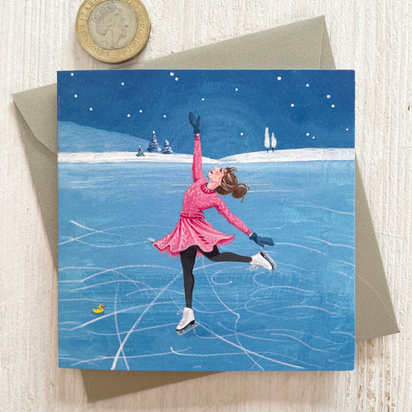 A square greeting card featuring an illustration of a woman ice skating on a blue frozen lake at night under a starry sky, with a silver pearlescent envelope behind it and a pound coin in the top-left corner for scale, on a light wood surface.