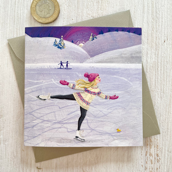 A square greeting card featuring an illustration of a woman ice skating on a frozen lake at nsunset, with a silver pearlescent envelope behind it and a pound coin in the top-left corner for scale, on a light wood surface.