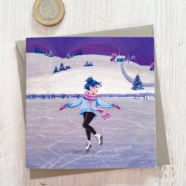 A square greeting card featuring an illustration of a woman ice skating on a frozen lake at night , with a silver pearlescent envelope behind it and a pound coin in the top-left corner for scale, on a light wood surface.