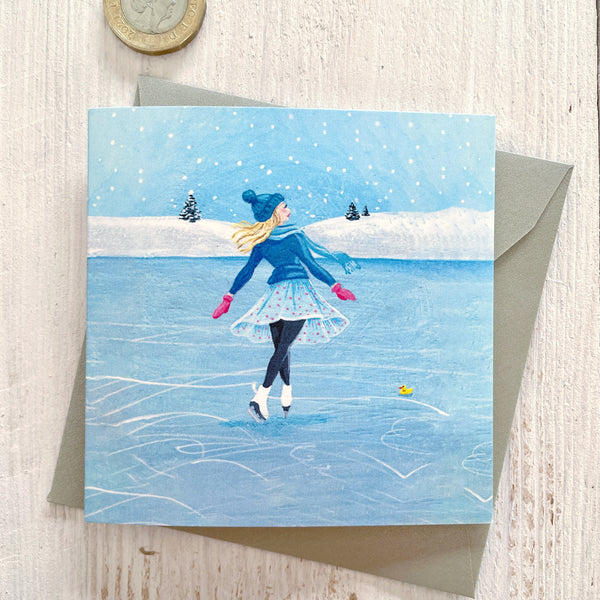 A square greeting card featuring an illustration of a woman ice skating on a blue frozen lakein the snow, with a silver pearlescent envelope behind it and a pound coin in the top-left corner for scale, on a light wood surface.