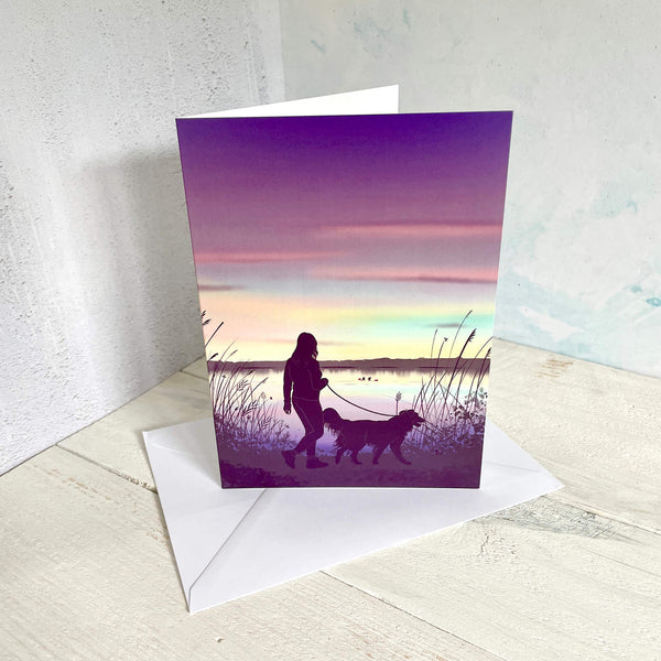 Greeting card with a silhouette of a person walking a dog at sunset, standing on a wooden surface with an envelope.