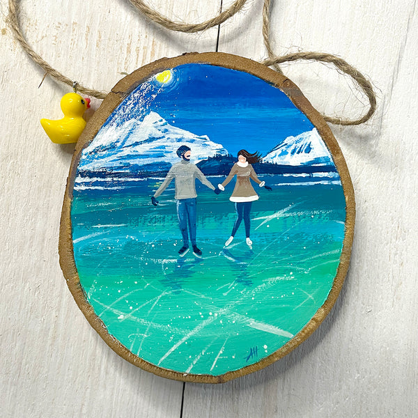 Hand-painted wood slice artwork showing two ice skaters holding hands on turquoise ice with snowy mountains in the background.