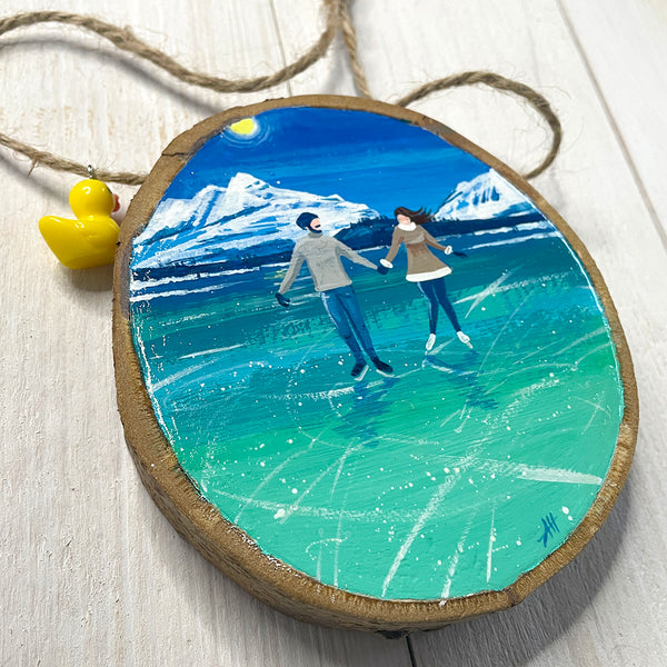 Hand-painted wood slice artwork showing two ice skaters holding hands on turquoise ice with snowy mountains in the background.