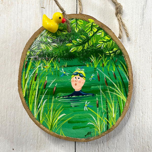 Hand-painted wood slice artwork showing a swimmer in green water surrounded by reeds and dragonflies beneath leafy branches