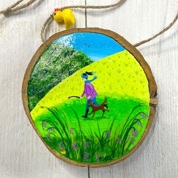 Hand-painted wood slice artwork showing a woman walking her dog across bright green countryside fields with purple wildflowers.