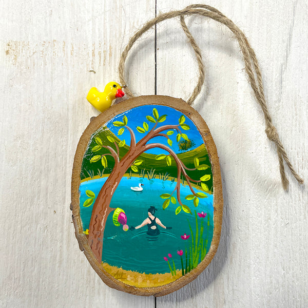 Hand-painted wood slice artwork showing a miniature wild swimmer in a calm pond beneath an overhanging tree with spring leaves and wildflowers.