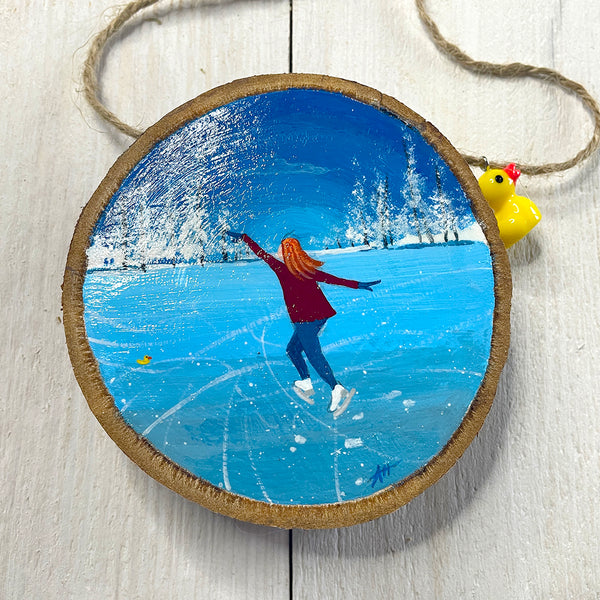 Hand-painted wood slice artwork showing a lone ice skater gliding across frozen blue ice with snowy trees in the background.