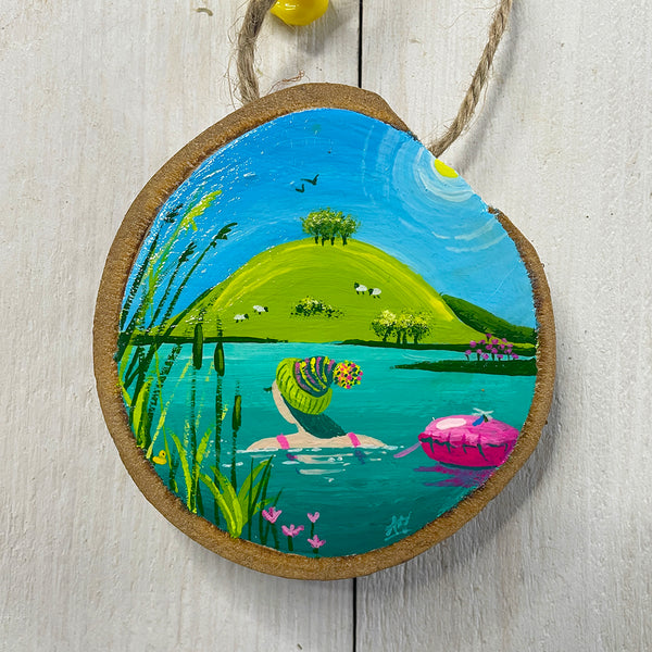 Hand-painted wood slice artwork showing a wild swimmer wearing a bobble hat with a bright pink tow float on calm blue water and sheep on a green hill.
