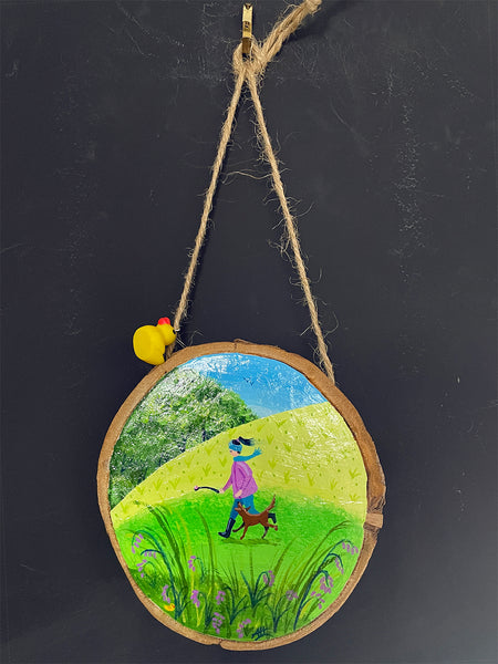 Original painted log slice decoration featuring a countryside walking scene hanging against a blackboard background.