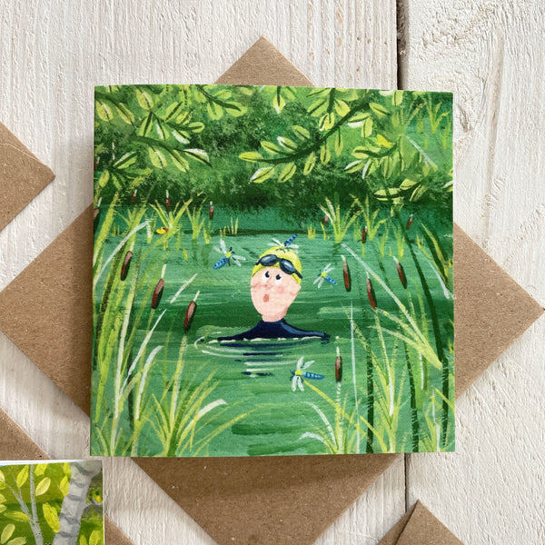 Card with a cswimmer in a pond surrounded by dragonflies and greenery, on a wooden surface.