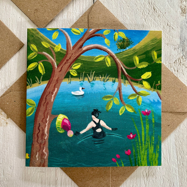Card with a painting of a person in a swimsuit entering a lake with a bobble hat hanging on a branch, placed on a wooden surface with a natural brown envelope.