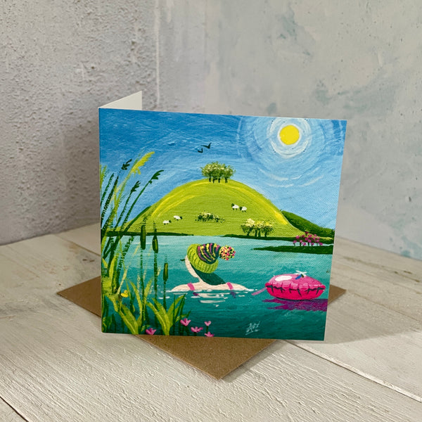 Greeting card with a scenic illustration of a lake, with a swimmer and tow float, placed on a wooden surface.