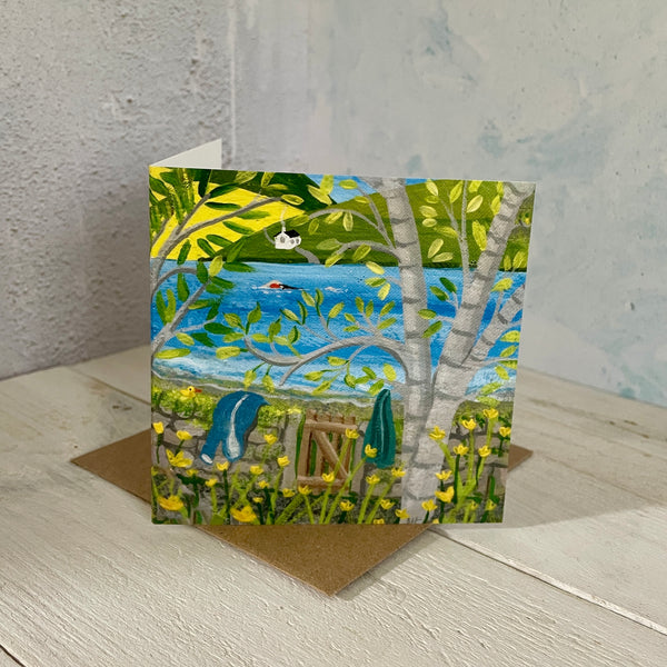 Greeting card with a nature scene featuring a gate, trees, and a lake with a swimmer, placed on a wooden surface.