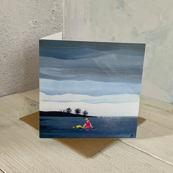 Mini greeting card with a painting of a swimmer in a lake, placed on a wooden surface.