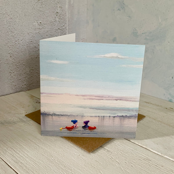 Greeting card with a painting of two swimmers with tow floats in a lake, placed on a wooden surface.