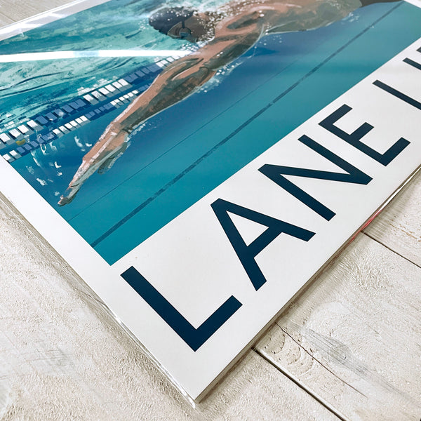 Lane Life swimming print photographed at an angle, showing the male swimmer, pool lane and bold typography in a cellophane bag