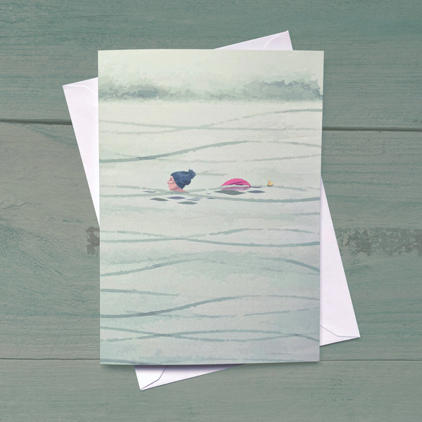 A greeting card with a serene swimming scene, featuring a person swimming in calm water, presented with a white envelope on a blue wooden background.