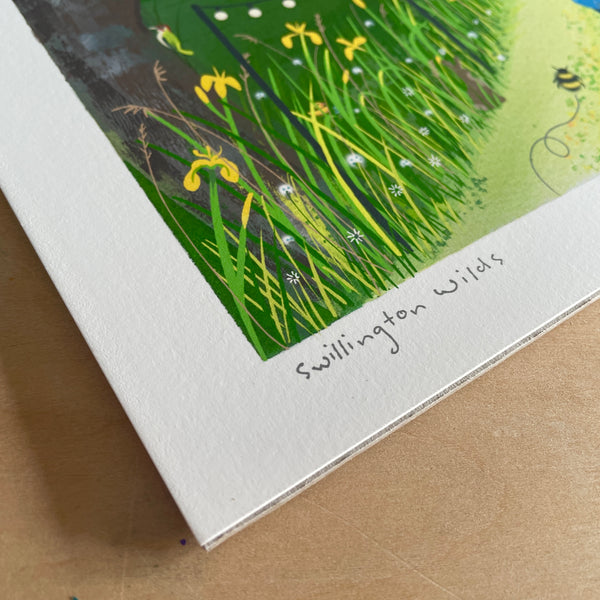 A close-up of a printed artwork showing the texture of the paper with a grassy lake edge with yellow Iris flowers, green leaves, tree trunk and a bee, with the handwritten text 'Swillington Wilds' along the white border. The print is photographed on a wooden surface.