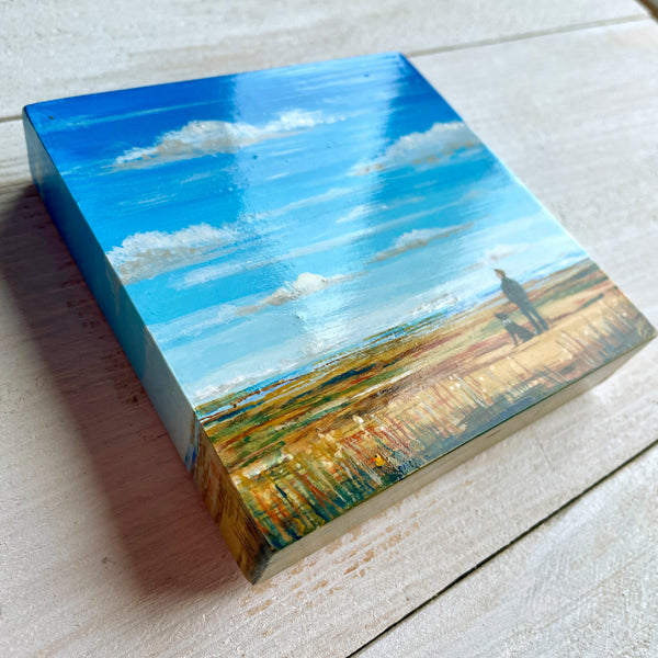 Small square painting of a landscape with a blue sky and green field on a wooden surface.