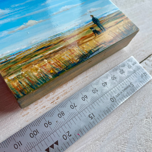 Small painting of a landscape with a person and dog, next to a ruler for scale.