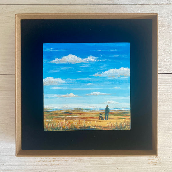 Boxed painting of a man and dog looking up at a big blue sky.
