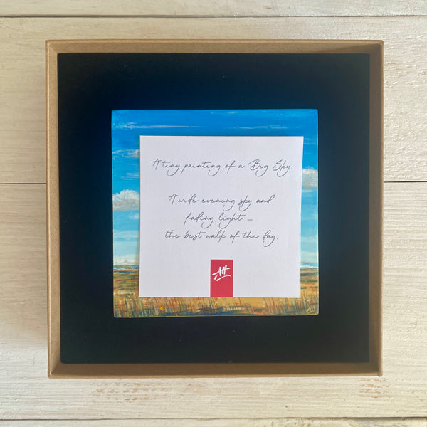 Boxed artwork with description card and a small painting on a wooden surface