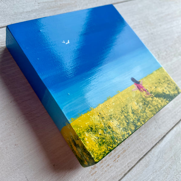 Original Miniature Big Sky painting of rapeseed fields beneath a blue sky with a figure walking through. Shown at an angle to show off the gloss varnish