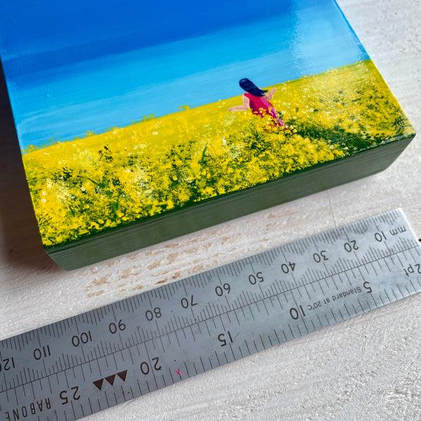 Crop of original miniature Big Sky painting of rapeseed fields beneath a blue sky with a figure walking through. Show with a ruler alongside for scale
