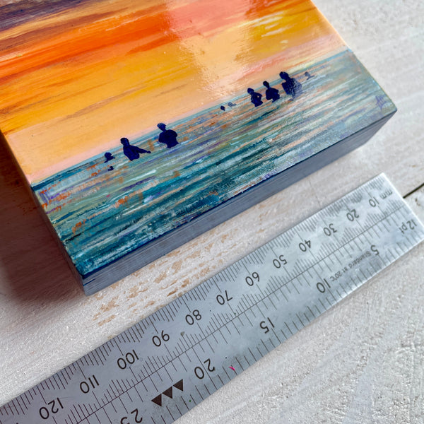 Small painting on a wooden panel of sea swimmers at sunset, next to a ruler for scale.