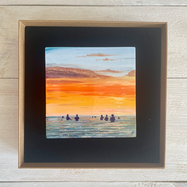 Boxed artwork of a sunset over water with silhouettes, on a wooden surface.