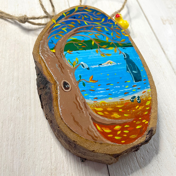 Hand-painted log slice artwork showing an autumn lakeside swim scene, with golden leaves falling and swimmers in blue water.
