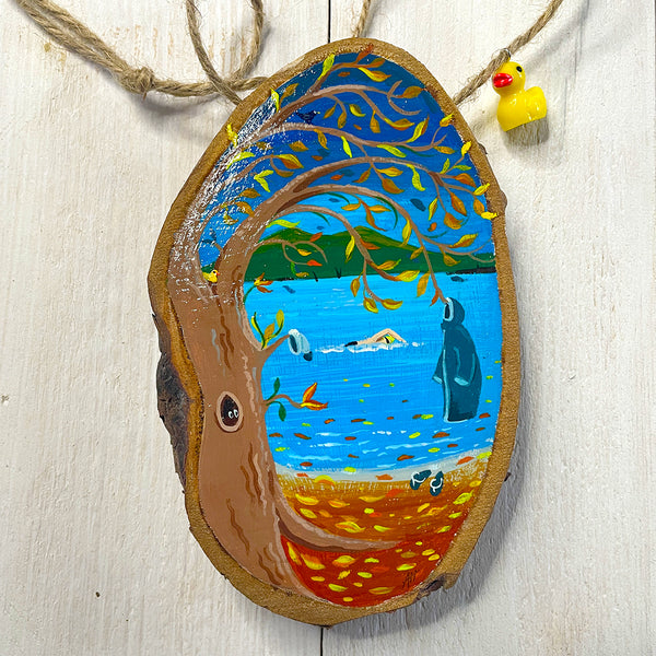 Hand-painted log slice artwork showing an autumn lakeside swim scene, with golden leaves falling and swimmers in blue water.