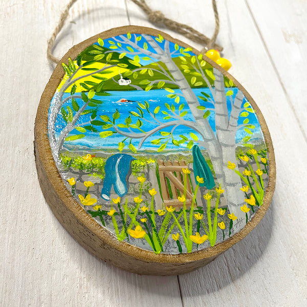 Hand-painted wood slice artwork showing a spring lakeside swim scene with birch trees, daffodils and a swimmer in blue water.