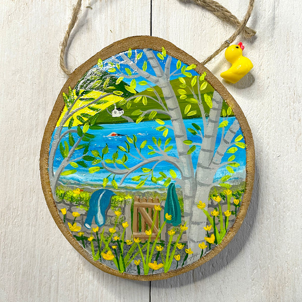 Hand-painted wood slice artwork showing a spring lakeside swim scene with birch trees, daffodils and a swimmer in blue water.