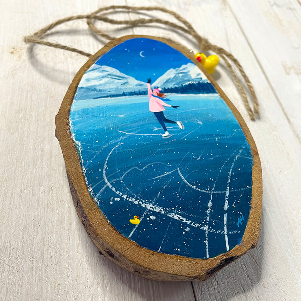 Hand-painted log slice artwork showing an ice skater on frozen blue ice with snowy mountains and falling snow.