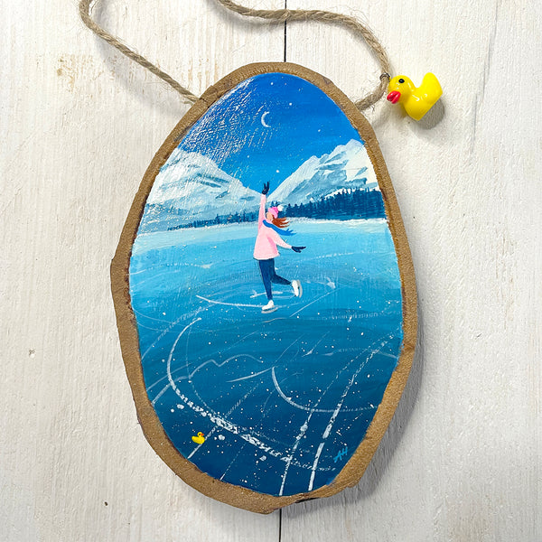 Hand-painted log slice artwork showing an ice skater on frozen blue ice with snowy mountains and falling snow.