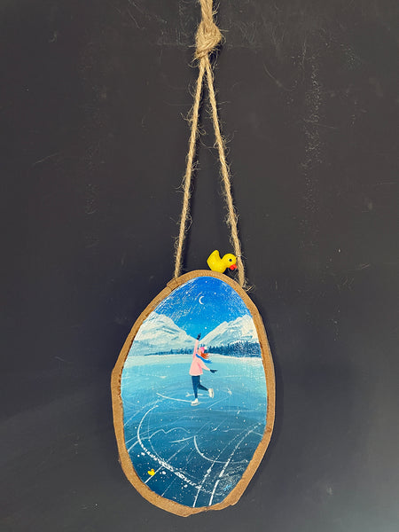 Original painted wood slice decoration featuring winter ice skating scene, hanging against with a Ducky charm against a blackboard background.